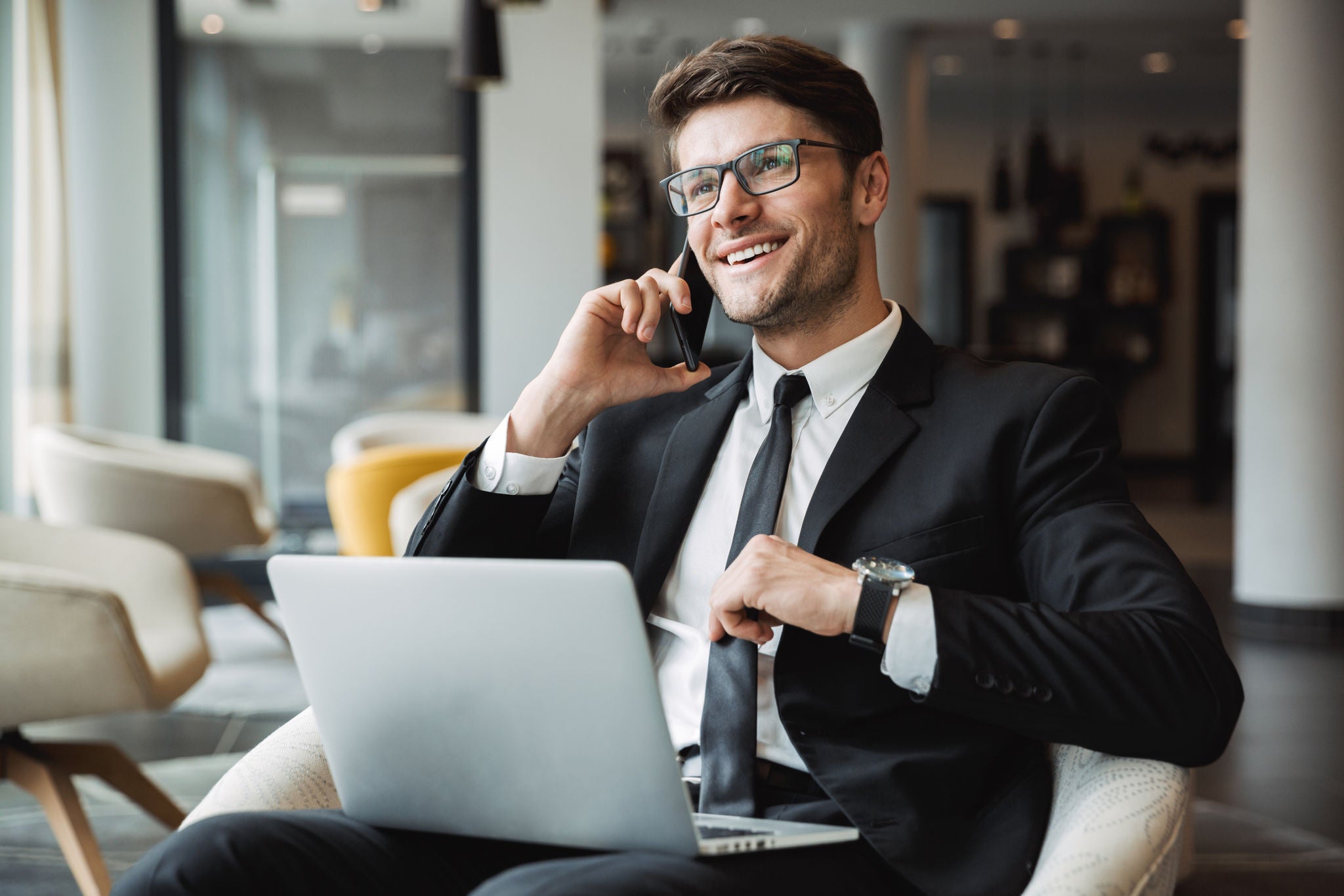 Portrait of happy young businessman wearing formal black suit using laptop computer and talking on smartphone in hotel hall